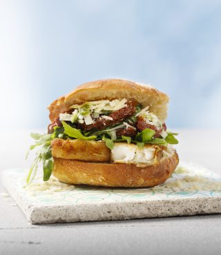 Ein Sandwich mit knusprigem FRoSTA-Fisch, Rucola, sonnengetrockneten Tomaten, geriebenem Käse und Soße auf einem knusprigen Brötchen, Steinfliesen, blauer Hintergrund.