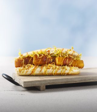 Ein Sandwich mit goldbraun frittierten FRoSTA-Sticks, gelbem Topping, gehackten Nüssen und cremiger Sauce auf Holzbrett vor hellem Hintergrund.