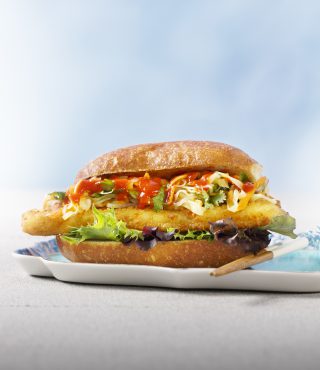 Ein knuspriges FRoSTA-Fischbrötchen mit Salat, Krautsalat und roter Sauce auf einem weichen Brötchen, serviert auf einem Teller vor hellblauem Hintergrund.