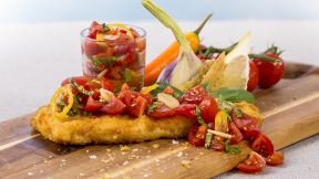 Ein knuspriges FRoSTA Fischfilet mit frischer Tomatensalsa wird auf einem Brett mit Zitrone, Basilikum, Knoblauch, Gemüse und extra Salsa serviert.