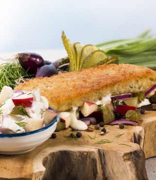 Ein paniertes FRoSTA Fischfilet-Sandwich mit Käse, roten Zwiebeln, Gurken und Kräutern. Dazu Salat, frische Kräuter und Dip nach Hausfrauenart.