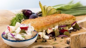 Ein paniertes Sandwich mit geschmolzenem Käse und Kräutern sowie FRoSTA-Tiefkühlprodukten liegt neben Apfelsalat und Dip auf Holzbrett.