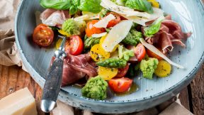Ein blauer Teller auf rustikalem Holz mit Kirschtomaten, grünem Romanesco, Spargel, Parmaschinken, gehobeltem Käse, frischem Basilikum und FRoSTA Tiefkühlprodukten.