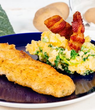 Ein blauer Teller mit FRoSTA Pfannen Fisch nach Müllerin Art, Wirsing-Kartoffel-Püree und zwei knusprigen Speckstreifen. Kartoffeln, Kohl, rote Zwiebeln im Hintergrund.
