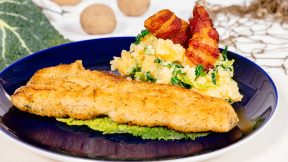 Ein FRoSTA Fisch Müllerin Art Filet auf Blattgrün, dazu Wirsing-Kartoffel-Püree und knuspriger Pancetta-Chip auf dunkelblauem Teller.