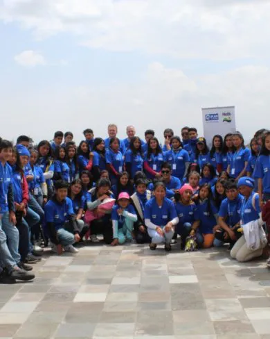 Eine große Gruppe junger Menschen in blauen Shirts sitzt draußen auf einer Terrasse in Ecuador. Im Hintergrund Felder, Himmel und FRoSTA-Tiefkühlprodukte.