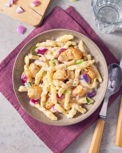 Eine Schüssel cremige Pasta mit FRoSTA Tiefkühlprodukten, gegrilltem Hähnchen, roten Zwiebeln und grüner Paprika – dazu Besteck und Wasserglas.