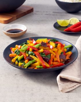 Ein schwarzer Teller gefüllt mit farbenfrohem FRoSTA-Wok-Gemüse – rote/gelbe Paprika, grüne Bohnen, Auberginen – plus Soße, Limettenspalten, Chili und einer Serviette daneben.