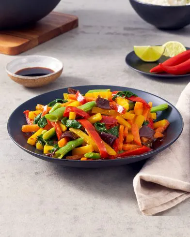 Ein schwarzer Teller gefüllt mit farbenfrohem FRoSTA-Wok-Gemüse – rote/gelbe Paprika, grüne Bohnen, Auberginen – plus Soße, Limettenspalten, Chili und einer Serviette daneben.