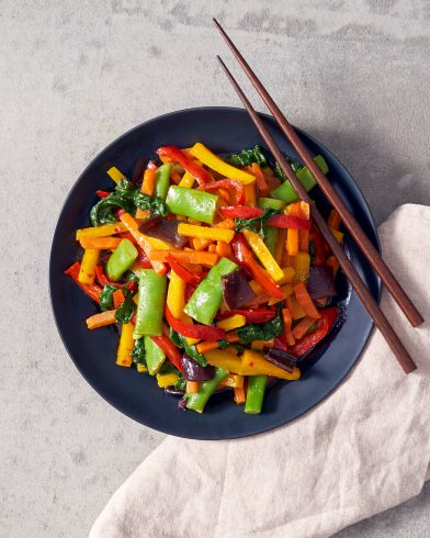 Neben Stäbchen und Serviette liegt ein schwarzer Teller mit lebendigen FRoSTA Tiefkühlprodukten wie Wokgemüse, Paprika, Erbsenschoten und Karotten.