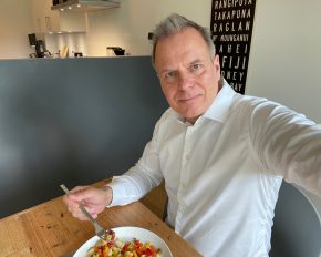 Ein Mann im weißen Hemd sitzt an einem Holztisch, genießt einen bunten Salat mit FRoSTA Tiefkühlprodukten und lächelt in seiner modernen Küche.