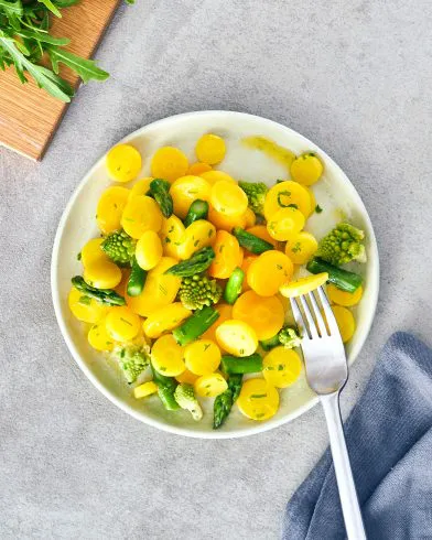 Ein Teller mit FRoSTA-Tiefkühlprodukten: gelbe Karotten, grüner Spargel und Romanesco, dazu Rucola, Gabel und graue Serviette.