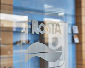 Eine Glastür mit dem Schriftzug FRoSTA und dem Wellenlogo. Verschwommenes blau-braunes Interieur, Regale, Wandobjekte und subtile Lichtreflexe im Hintergrund.