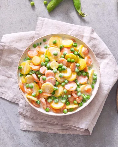 Eine Schüssel cremiges FRoSTA-Mischgemüse – Karotten, Kartoffeln, grüne Erbsen – ist die perfekte FRoSTA-Gemüsebeilage auf beigem Tuch.