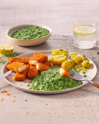 Ein Teller mit panierten FRoSTA Fischstäbchen, FRoSTA Rahmspinat und Salzkartoffeln mit Kräutern. Ein Glas Zitronenwasser, im Hintergrund eine Schale mit extra FRoSTA Rahmspinat.
