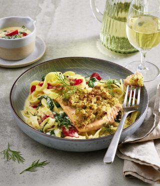 Ein Teller FRoSTA Wildlachs Tagliatelle mit Spinat und Paprika, belegt mit Kräuter-Lachsfilet. Daneben Weißwein, Krug und Spinatsoße.