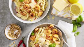 Zwei Schüsseln cremige Pasta mit Garnelen von FRoSTA, getoppt mit Basilikum und Parmesan, umgeben von Zitrone, Käseblock, Pinienkernen und Chili.