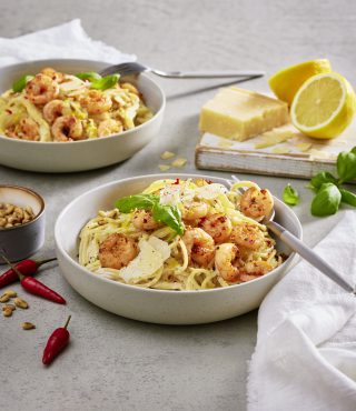 Zwei Schüsseln Pasta mit FRoSTA Garnelen, geriebenem Käse und Basilikum stehen bereit, dazu Nüsse, Chili, Zitrone, Käseblock und frisches Basilikum.