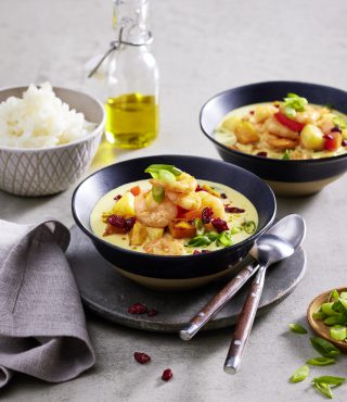 Zwei Schüsseln cremiges FRoSTA Garnelen Curry mit Gemüse, garniert mit grünen Zwiebeln, stehen neben Reis, Öl und Cranberries auf dem Tisch.