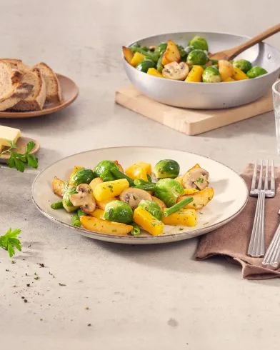 Ein Teller FRoSTA Gemüsepfanne mit Kartoffeln, Rosenkohl, gelbem Paprika und Champignons steht neben Wasser, Brot und Besteck.