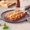 Serviert wird ein Stück FRoSTA Lasagne Bolognese mit Fleischsauce und Käse, garniert mit Basilikum, im Hintergrund sind Käsewürfel und Tomaten.