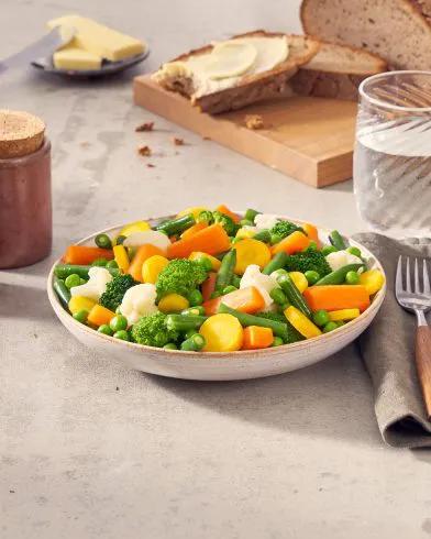 Ein Teller mit FRoSTA Gemüsemix – gedünstete Karotten, Brokkoli, Bohnen, Erbsen und Blumenkohl – daneben Wasser, Besteck, Serviette und Butterbrot.