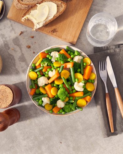 Eine Schale FRoSTA-Gemüsemix – Karotten, grüne Bohnen, Brokkoli, Blumenkohl und Erbsen – steht mit Brot und Wasser auf dem Tisch.