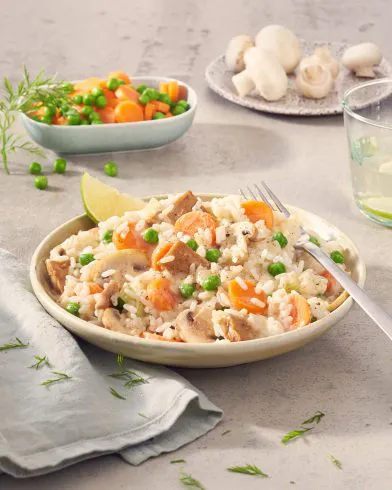 Ein Teller FRoSTA Veganes Hühnerfrikassee mit Reis, Erbsen, Karotten und Pilzen steht neben Besteck und frischem Gemüse auf dem Tisch.