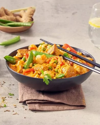 Eine schwarze Schüssel mit FRoSTA Tiefkühlprodukten: buntem Gemüse, Tofu und Reis im roten Curry steht auf einer braunen Serviette mit Stäbchen.