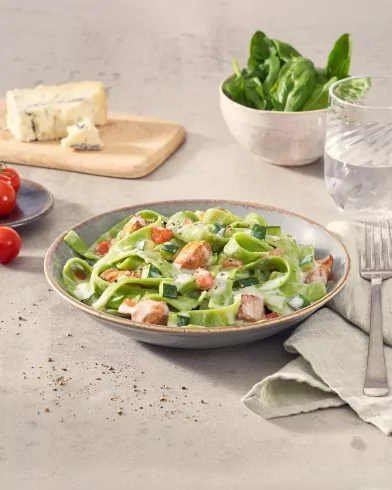 Eine Schüssel cremige Tagliatelle Rahm Hähnchen FRoSTA mit Spinat und gewürfeltem Gemüse steht neben Wasser, Spinat, Kirschtomaten und Käse.