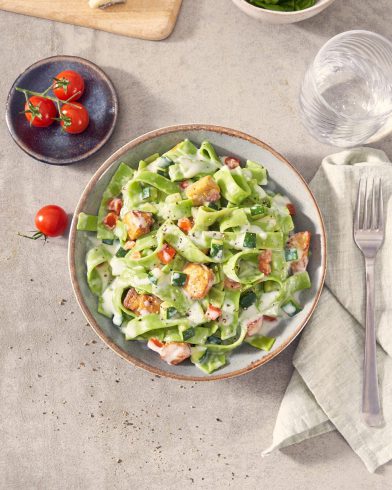 Eine Schüssel FRoSTA green Tagliatelle Rahm Hähnchen mit cremiger Sauce, Gemüse und Hähnchen steht auf dem Tisch; Dazu Besteck, Wasser und Käse.