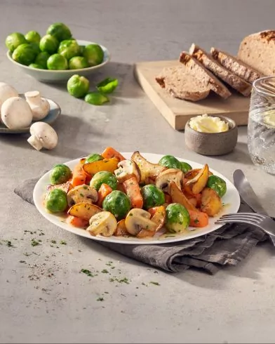 Ein Teller mit gekochtem FRoSTA Gemüse traditionell – Rosenkohl, Karotten, Pilzen, Kartoffeln – mit Brot, Butter und Wasser auf dem Tisch.