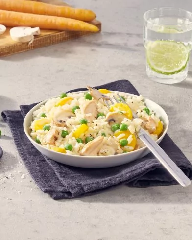 Eine Schüssel cremiges Risotto mit FRoSTA Erbsen, gelben Paprikaschoten, Champignons und zartem Hähnchenfleisch steht auf einer dunklen Serviette. Gabel in der Schüssel.