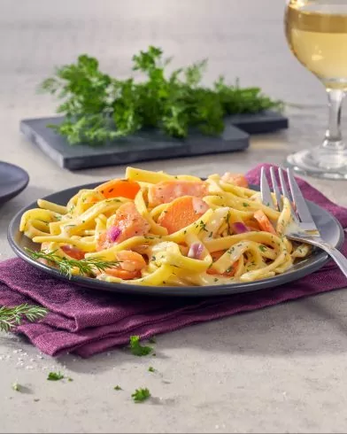 Ein Teller cremige Pasta mit FRoSTA Wildlachs, Karotten, roten Zwiebeln und Kräutern wird auf einem grauen Teller mit einer Gabel und einer lila Serviette serviert.