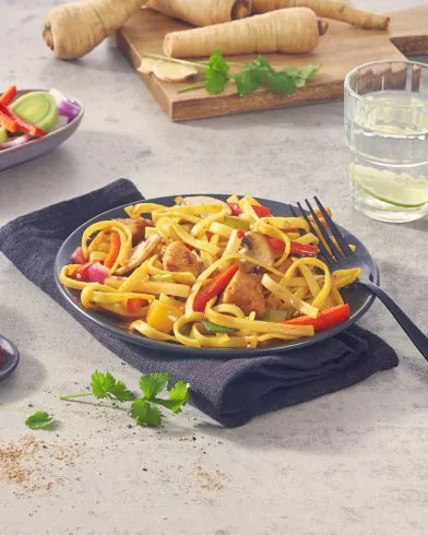 Ein Teller mit FRoSTA Woknudeln mit Gemüse und Tofu steht auf einer Serviette neben einer Gabel, im Hintergrund stehen Wasser, Pastinaken und Salat.