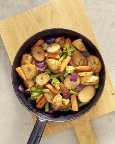 Eine Bratpfanne mit einer bunten FRoSTA Bratkartoffelpfanne aus Kartoffelscheiben, Karotten, grünen Bohnen, Brokkoli und roten Zwiebeln auf einem Brett.