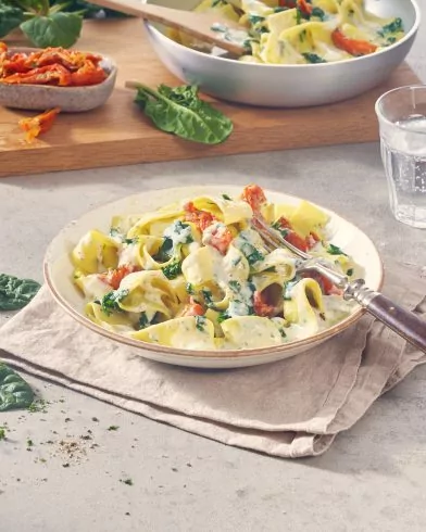 Ein Teller cremige Pappardelle mit FRoSTA-Tiefkühlspinat und getrockneten Tomaten liegt neben Gabel und Wasserglas auf einer beigen Serviette.