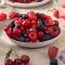 Eine weiße Schale mit frischen Beeren und Kirschen, ideal zum Genießen oder für FRoSTA tiefgekühlte Leckereien, steht mit weiteren Beeren und Eiswasser auf einem Tisch.