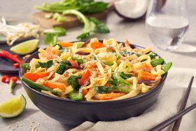 Eine schwarze Schüssel mit bunten FRoSTA Gemüsepfannennudeln, Sesamkörnern und frischem Grün steht auf einer beigen Serviette – daneben Limette und Essstäbchen.