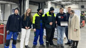 Sieben Personen stehen an einem Weltmädchentag lächelnd mit Laptop und FRoSTA-Tiefkühlprodukten in einer modernen Industriehalle zusammen.