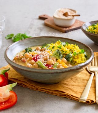 Eine Schüssel FRoSTA Gemüsecurry mit Kichererbsen, Brokkoli und Paprika, serviert mit gelbem Reis, goldenem Besteck, Wasser und Tomaten.