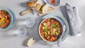 Zwei Schüsseln Nudel- und Gemüsesuppe, inspiriert von klassischer Minestrone mit FRoSTA Tiefkühlprodukten, dazu Brot und Wasser auf hellem Tisch.