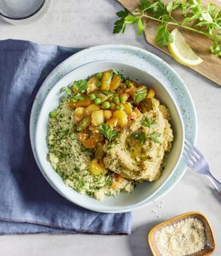 Eine vegane Bowl mit FRoSTA-Tiefkühlprodukten wie Couscous, Gemüse, Erbsen und Kräutern auf blauem Tuch mit Beilagen serviert.
