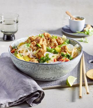 Eine Schüssel mit FRoSTA-Tiefkühlprodukten, Reisnudeln, Gemüse und frischen Kräutern, serviert mit Erdnussdressing und Limettenstück.