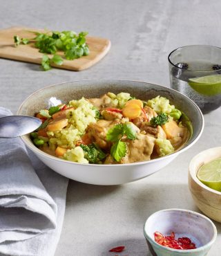 Eine Schüssel FRoSTA Hähnchen Curry Rezept mit Reis, Gemüse und Kräutern steht neben Wasser mit Limette und Beilagen auf dem Tisch.