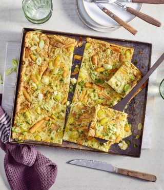 Eine gebackene Gemüsefrittata mit FRoSTA Tiefkühlprodukten und Orzo, in Quadrate geschnitten, mit Frühlingszwiebeln garniert. Ein Stück mit dem Spatel herausheben.