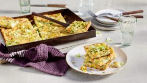 Ein Teller mit FRoSTA-Tiefkühlprodukt-Orzo-Pie, garniert mit Lauchzwiebeln, neben Blech mit mehr Stücken und Gläsern Wasser am Tisch.