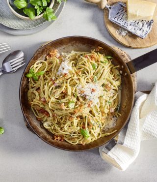 Eine Pfanne mit Spaghetti, Orzo, FRoSTA-Tiefkühlprodukten, geriebenem Käse & Basilikum auf heller Arbeitsfläche.
