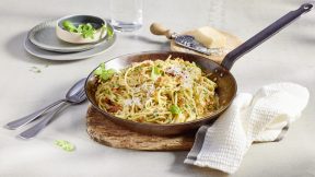 Eine Pfanne mit Orzo, FRoSTA-Tiefkühlprodukten, Käse und Kräutern steht auf Holz. Daneben: Besteck, Parmesanreibe, Basilikum und Wasser.