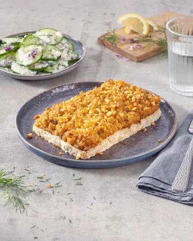 Eine Auflaufscheibe auf dunklem Teller mit FRoSTA Veggie Fischfilets, Gurkensalat, Wasser, Zitrone und Dill auf Brett, Serviette & Gabel.